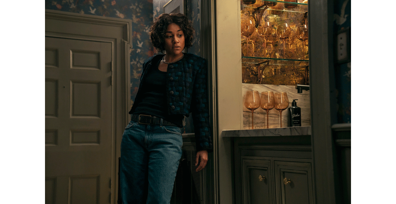 ジーンズ姿の巻き毛の女性が花柄の壁紙の壁にもたれている。女性がもたれた壁の横にはワイングラスが多数並んだ棚が見える。