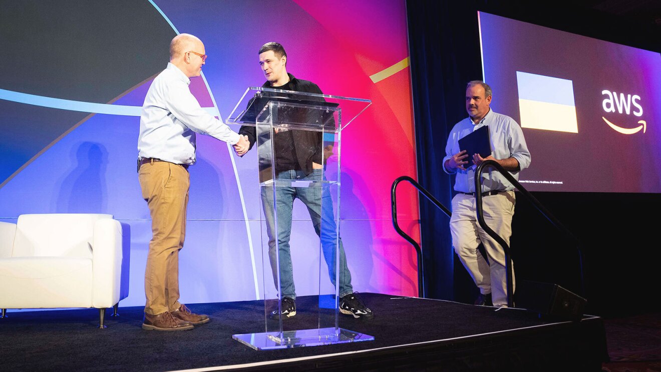 Two men shake hands on stage while another man walks to join them.