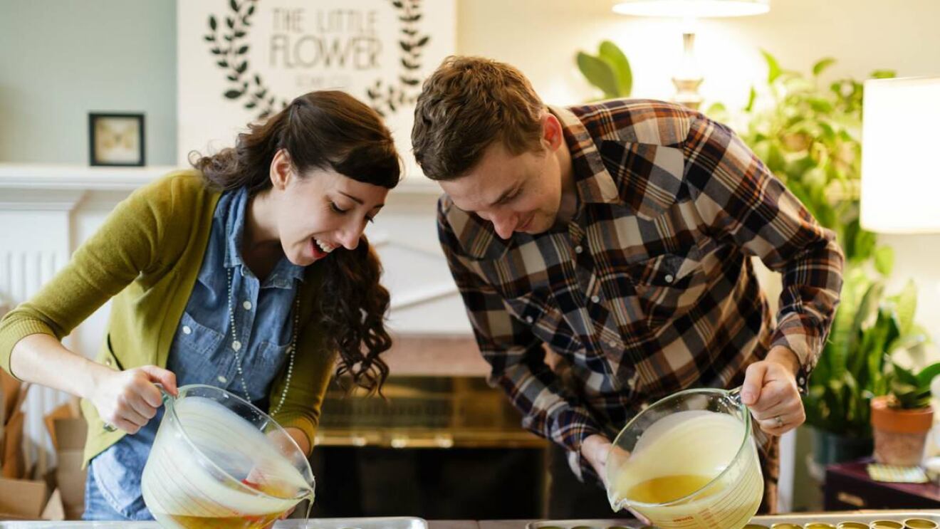 A woman (left) and man (right) hold large measuring cups filled with a liquid and pour the contents into small, metal containers. 