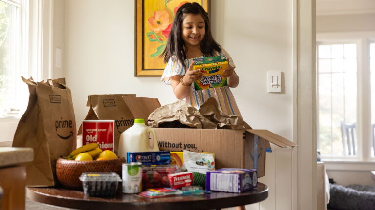 Girl unpacking Amazon Prime delivery box filled with groceries and household items on dining table