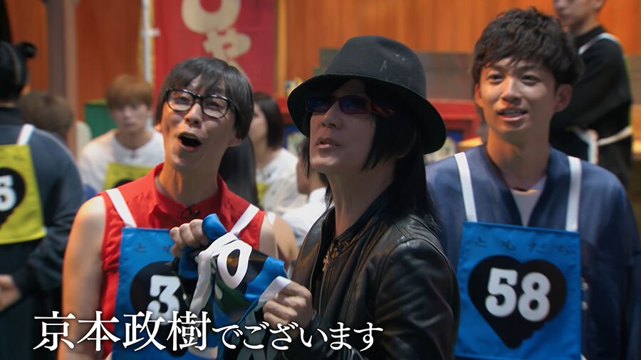 黒い帽子に黒い革ジャン、黒いサングラスの男性が青いゼッケンを持ちながら斜め上を見上げている。その横に青いゼッケンをつけた男性2人