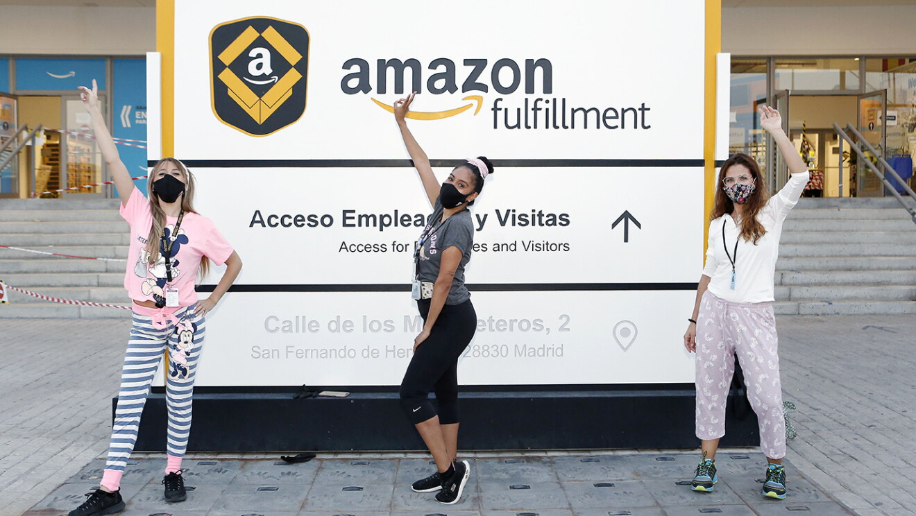 PJammin en MAD4, San Fernando de Henares. En la puerta principal de entrada al centro logístico hay tres asociadas posando delante del cartel Amazon fulfillment. Las tres tienen un brazo levantado. L aprimera va con un pijama de Mickey Mouse, rosa la parte superior y rayado el pantalón de color gris y balnco. La del medio va con un pantalón negro y la parte superior gris. La tercera lleva una parte superior de color blanco y una parte inferior rosa palido. Las tres van con deportivas, llevan mascasrilla. La primera y la tercera con el pelo largo suelto y la del meio con un moño alto.  