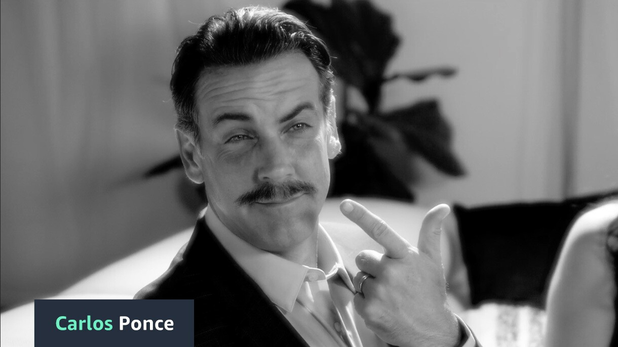 A black and white image of Carlos Ponce pointing at his mustache. The text in the bottom left corner reads "Carlos Ponce."