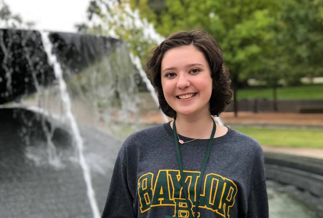 A young woman wearing a college sweatshirt stands in front of a fountain.