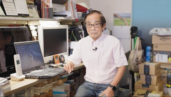 事務所兼倉庫のような室内で、椅子に座る男性。デスクにはノートパソコンやモニターがおこあれている。