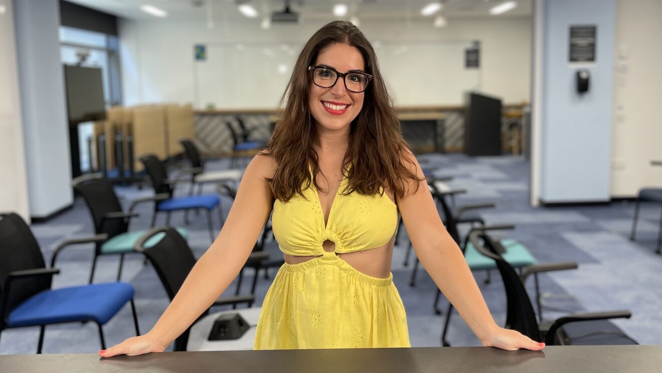 Maria Viñambres mirando a cámara con los manos apoyadas en la mesa de color gris. María tiene el pelo castaño ondulado y suelto, las uñas rosas, unas gafas de pasta y un vestido de color amarillo. De fondo se ve una sala con sillas y mesas de tonos azules y verdes y una pizarra blanca de fondo.