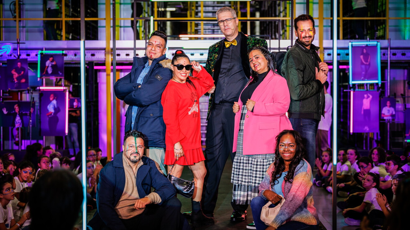 Dentro de un centro logístico, con las estanterías de fondo y las barandillas de color amarillo, aparece una pasarela de color negro ambientada con luces de neón y fotos de trabajadores de Amazon posando. Hay siete personas posando para la foto y mirando a cámara. Sentados a los dos extremos del grupo Óscar Urbano y Nazmilly Mosquera. El primero con un look de zapatos marrones, jersey beige, gabardina y pantalón azul marino. Lleva el pelo corto y barba y perilla. Y Nazmilly con sudadera multicolor, crop top azul, sus propios jeans y bolso a juego. Es una mujer racializada y lleva el pelo largo ondulado. De pie y de izquierda a derecha:  Christopher Rivadeneira con anorak con capucha, pantalón beige, deportivas azules y un toque personal de sus calcetines favoritos de Mario Bros. Se presenta con el pelo corto y los brazos cruzados. Lilimel Rivas lleva un vestido rojo, botas militares, bolso de mano y gafas cat eye. Íngrid Benítez, con el pelo largo y liso, luce una falda de cuadros vichy, americana rosa, zapatillas noventeras y bolso a conjunto con la americana. Juan Antonio Muñoz va con camisa y pantalón negro, pajarita de color amarillo y chaqué de lentejuelas de color verde. Y por último, Edgar Escobar, de lado mirando a cámara y un abrigo de plumas color caqui con camiseta amarilla, pantalones vaqueros y botas otoñales. Todos llevan la cara maquilladas con pinceladas de los colores de la bandera de su país.