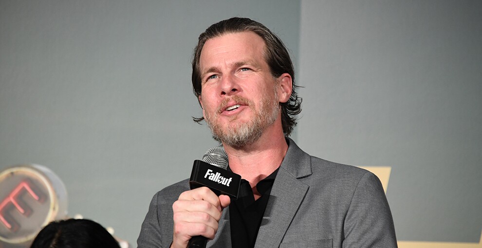 グレーの壁の前でマイクを持って話す男性。グレーのスーツに黒いシャツを着用。