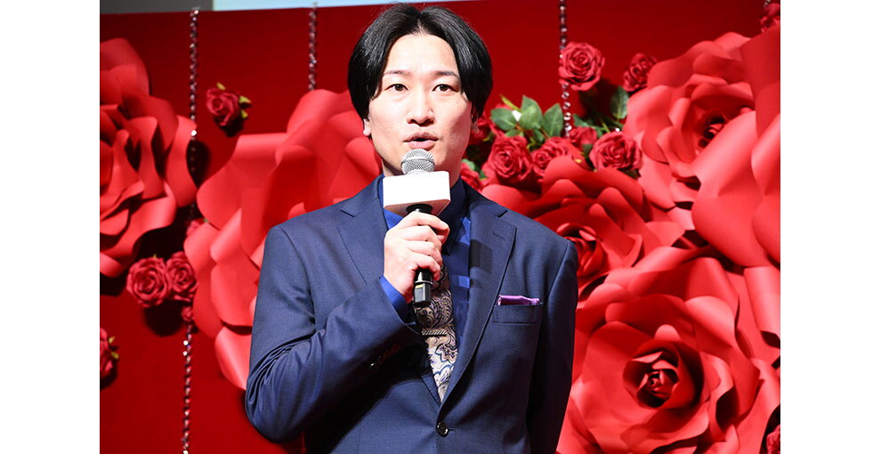 赤いバラの花の大きなオブジェと生花の赤いバラが飾られた赤い壁を背に、マイクを持って話すスーツ姿の男性。