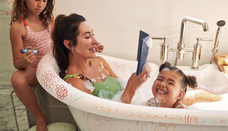 Frau liest auf ihrem Kindle mit ihren Kindern daneben, die in der Badewanne spielen.