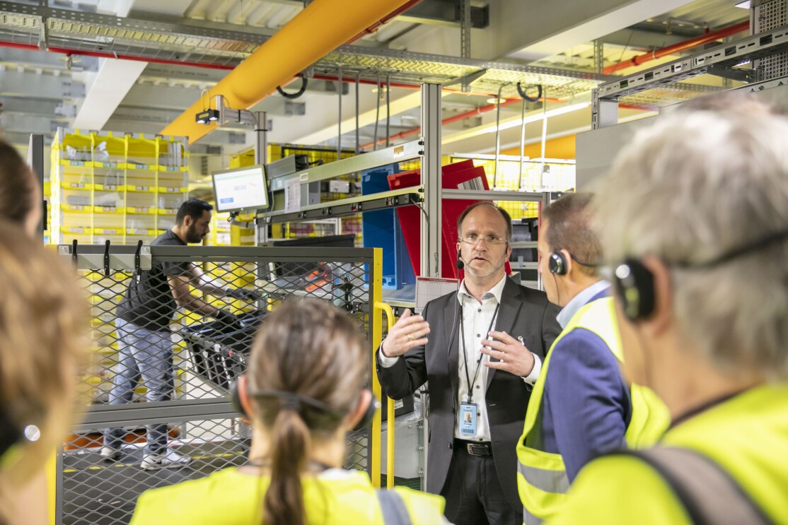 Ein Herr im Anzug steht vor einer Arbeitsstation im Logistikzentrum und erklärt Besuchern in gelben Besucherwesten den Ablauf
