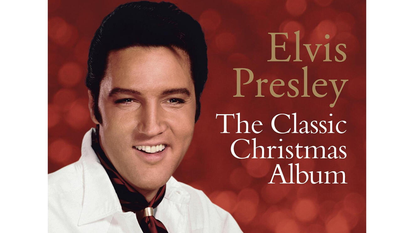 Elvis Presley wears a white button down and burgundy neck kerchief with a gold clasp. Behind him, we see out-of-focus lighting against a red background. 
