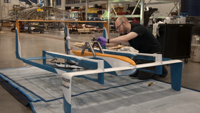 Amazon Prime Air drone in foreground. Behind the drone, a man is kneeling as he appears to be working on the drone. Near the drone, tools are seen.