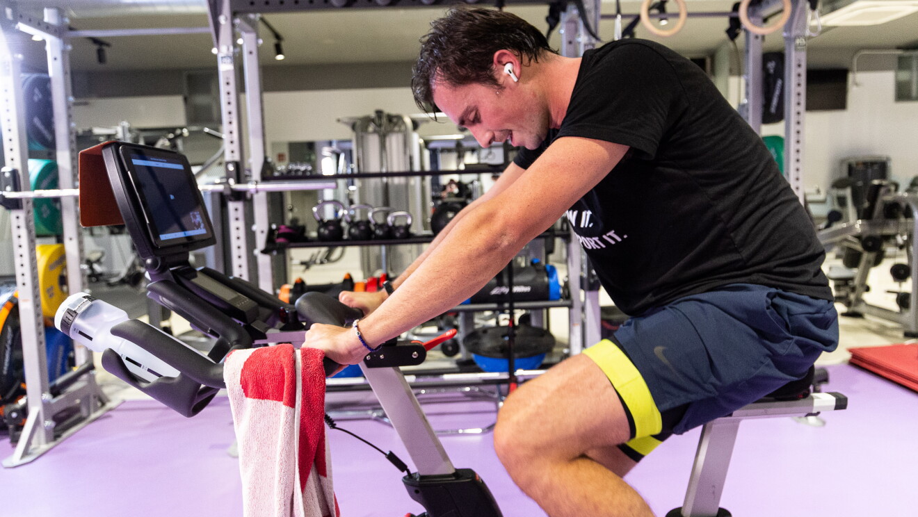 Tiago Moreira-Rato pedaleando para cumplir el reto. En un gimnaso está Tiago montado un bicicleta estática y escuachando música, Tiene un toalla apoyada en el manillar. 