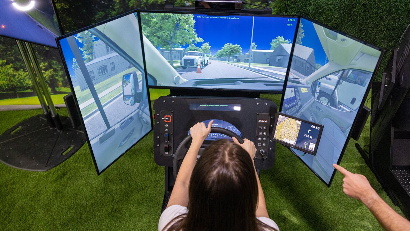 An image of a woman sitting in the driver's seat of an automated driving system. A person behind her is pointing to objects on the screen explaining what she is seeing.