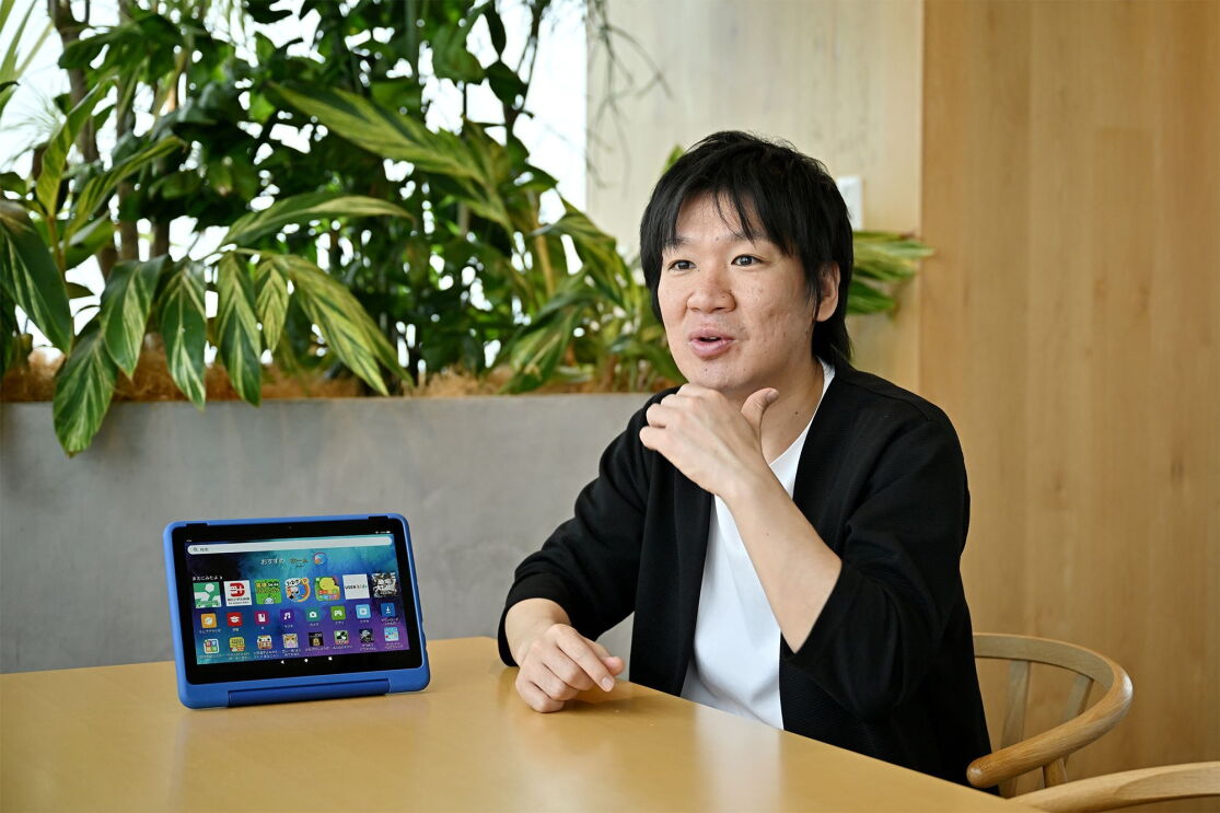 黒いジャケットを着た男性が顎に手をやり、話している。テーブルの上に青いタブレット