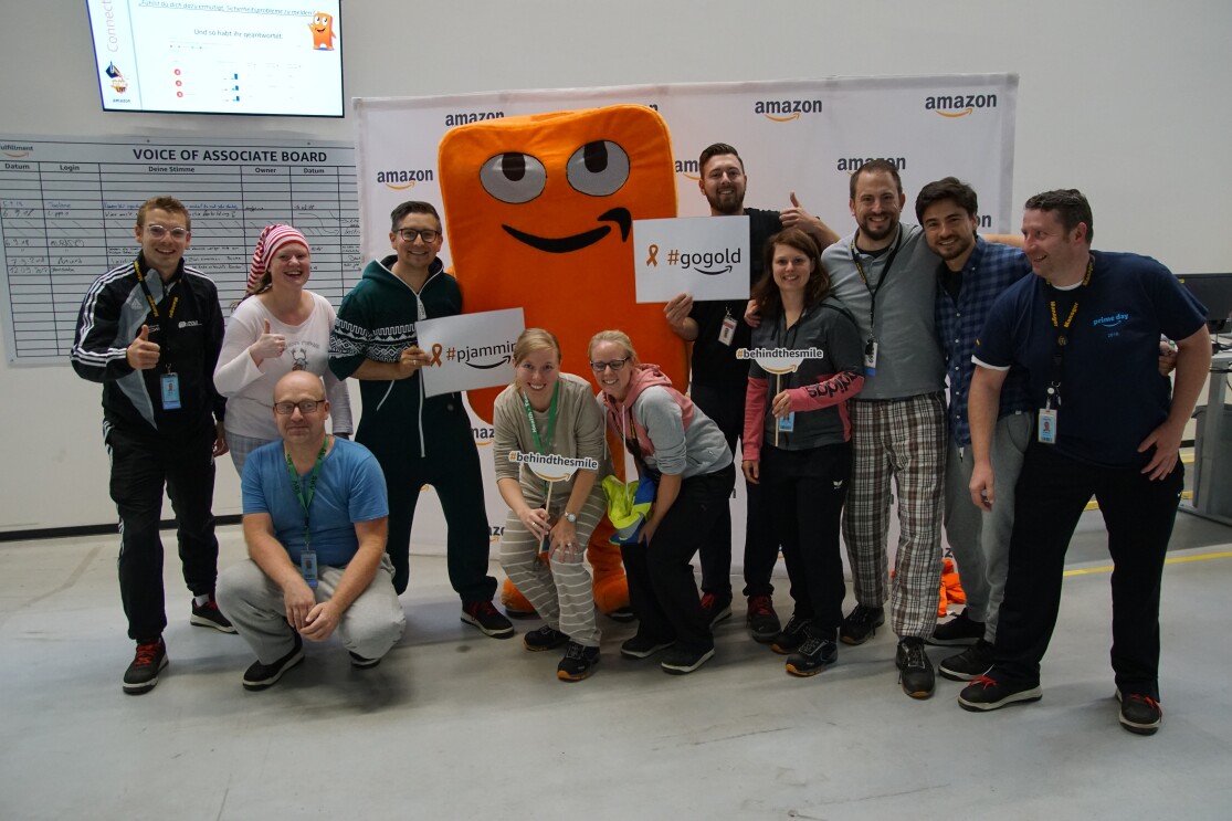 Eine Gruppe von Mitarbeiter steht vor einer Fotorückwand, in ihrer Mitte: das orangene Maskottchen Peccy in Form eines Amazon Pakets