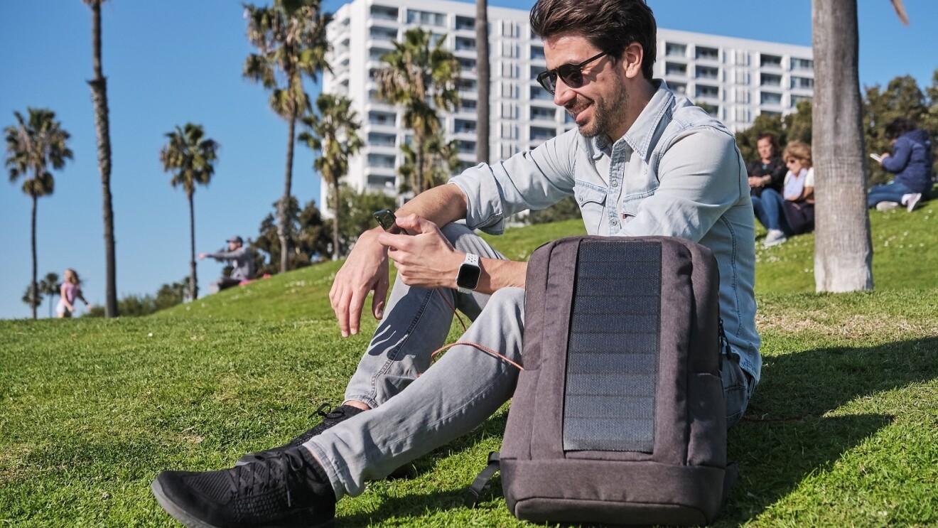 Ein Mann sitzt mit seinem Handy und einem Sunnybag, einem klimaschonend hergestellten Rucksack mit integrierten Solarpaneelen, auf einer grünen Wiese in der Sonne.