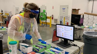 一名穿着全套COVID-19个人防护服和安全背心的妇女站在一张桌子旁边,桌子上有COVID-19检测用品和一台电脑显示器。