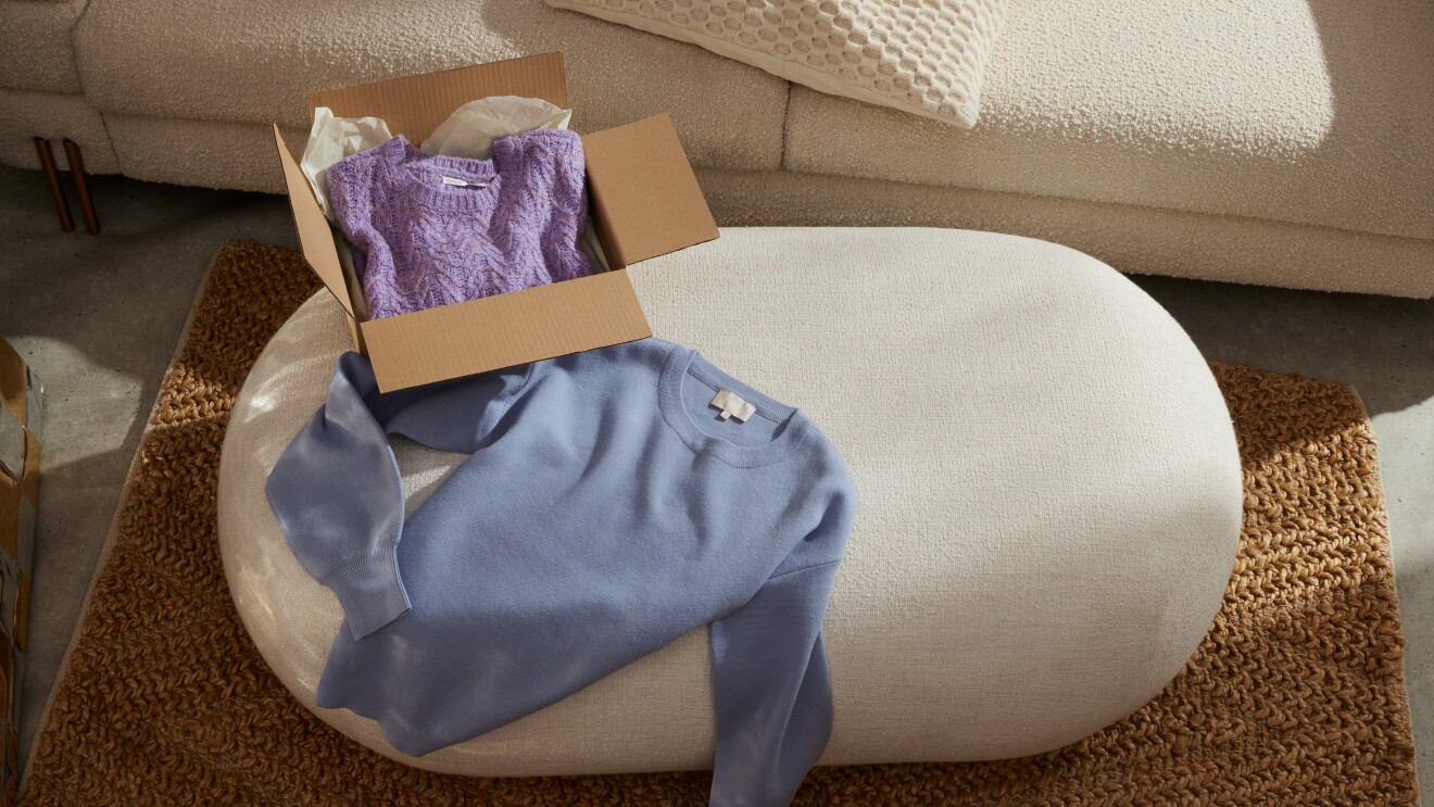 Immagine di un pouf sul quale è poggiato un pullover celeste e una scatola all'interno della quale si vede un altro pullover, questa volta lilla.