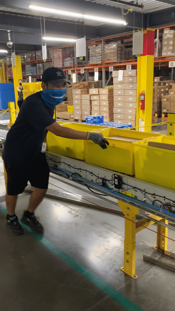 A man pushes yellow totes along a conveyor belt, then waves