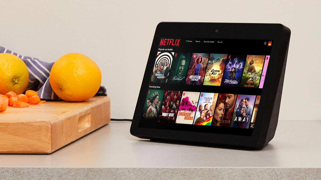 A black Echo Show sits atop a kitchen counter and displays a library of popular shows on Netflix. To the left of the device sits a cutting board with fruits and vegetables.