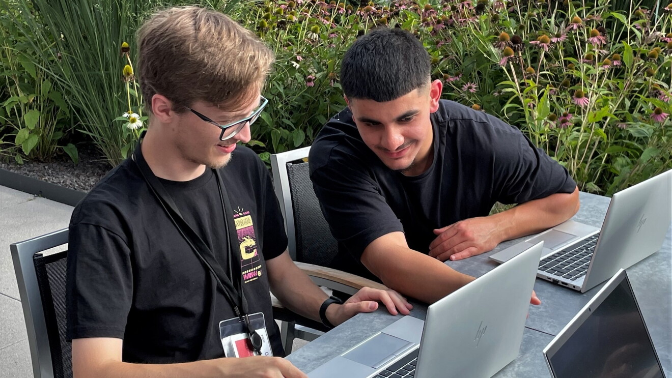 Die zwei AWS Azubis Hendrik und Mohammed sitzen draußen vor ihren Laptops und lächeln.