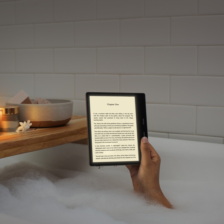 A woman holds a Kindle Oasis device with the page of a book on the display. Her arm appears from bubbles in a tub, with a wooden tub caddy behind the device.