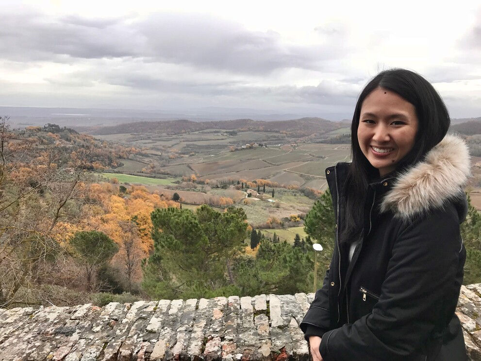 An image of a woman smiling for a photo in front of a scenic hillside. The trees are changing color with autumn. 