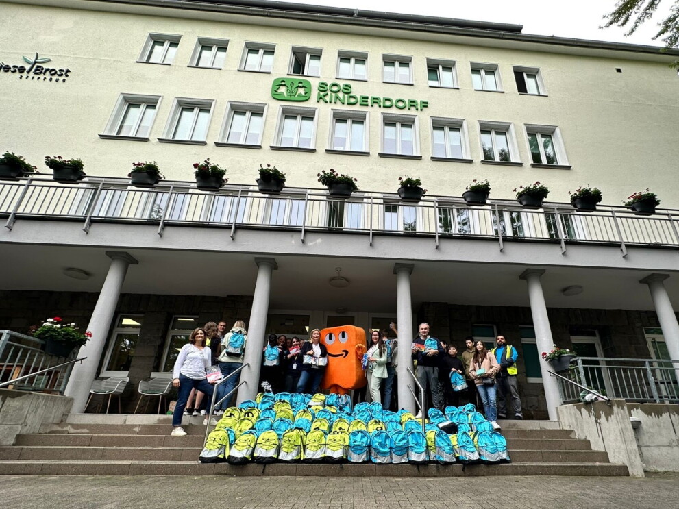 Amazon Mitarbeiterinnen und Mitarbeiter und Maskottchen Peccy stehen vor bunten Back to School Rucksäcken vor einem SOS Kinderdorf in Essen.