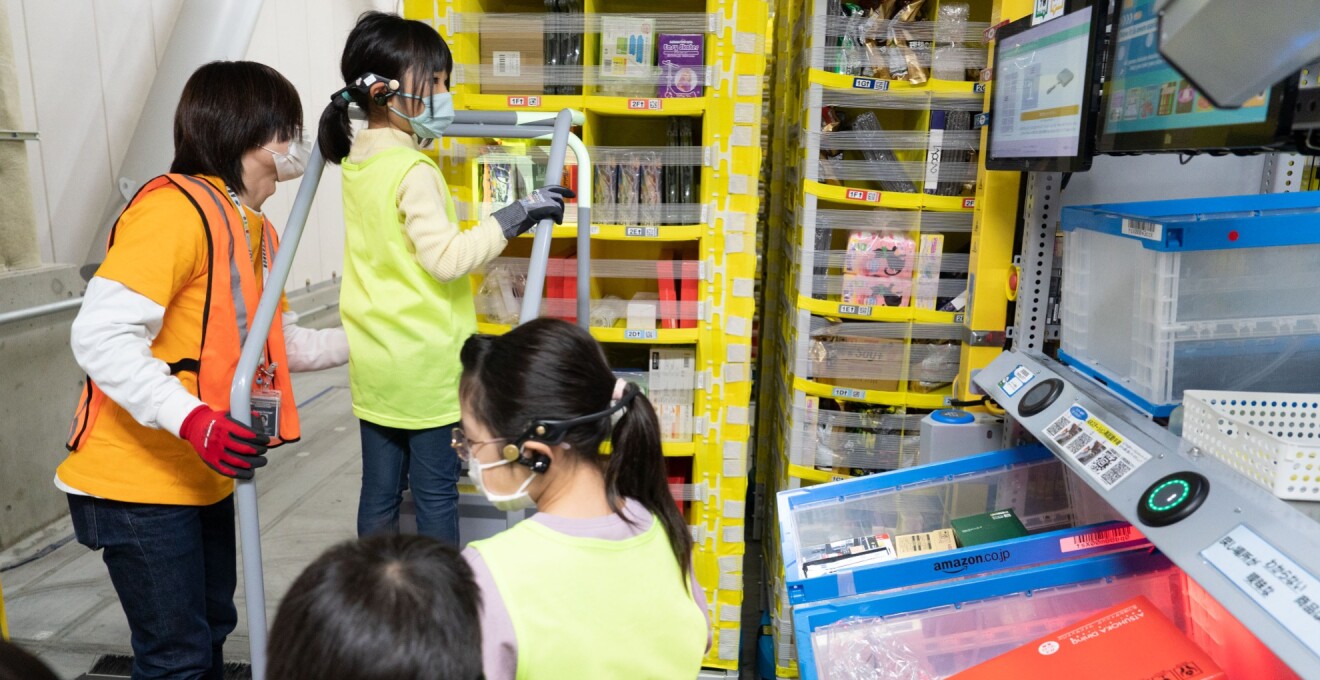 ロボットが運んできた消費棚の前で台に乗っている女の子とそれをサポートするAmazonのスタッフ