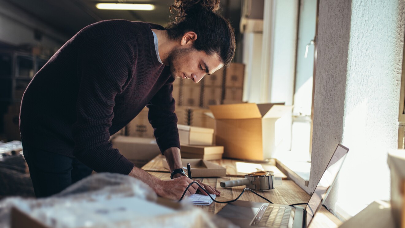 EIn Mann beugt sich über einen Schreibtisch, vor sich einen aufgeklappten Laptop, während im Hintergrund mehrere geöffnete Kartons zu sehen sind.