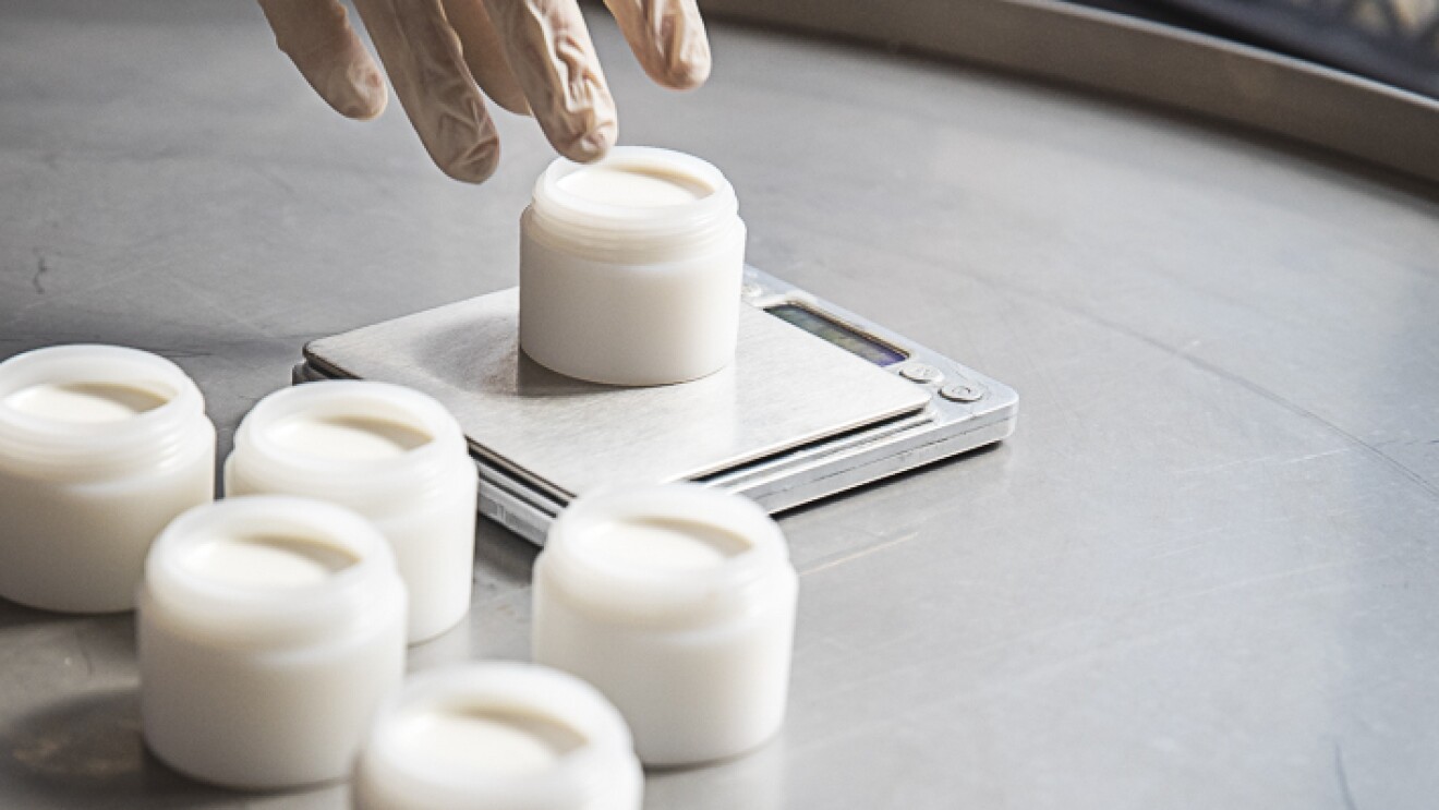 Auf einer grauen Arbeitsfläche stehen fünf geöffnete weiße Döschen, in die die handgemachte Deocreme Sommerliebe abgefüllt wurde. Am oberen linken Bildrand ist eine Hand mit Gummihandschuh zu sehen, die gerade ein sechstes Döschen auf eine Feinwaage gestellt hat.