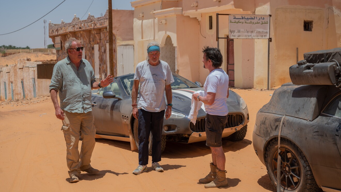 Three older men stand around a sports car 