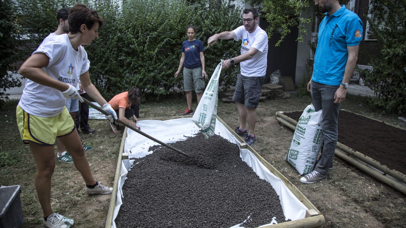 A scuola di verde: sei volontari Amazon all'opera nel giardino della scuola Media Leonardo da Vinci di Affori, nella periferia di Milano. Una donna in primo piano regge una pala con cui stende della terra, versata da un sacco da un uomo, in piedi di fronte alla donna. Gli altri 4 volontari osservano la scena.