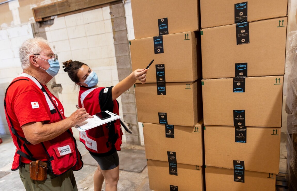 Two Red Cross volunteers stand next to two stacks of Amazon boxes filled with relief items.