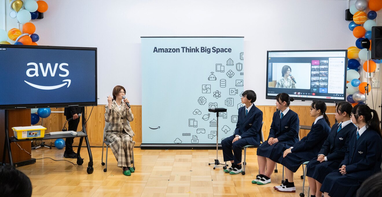 ベージュのチェック柄スーツを着た発表者が、紺色の制服姿の生徒たちの前でマイクを持って座って話している。会場には「Amazon Think Big Space」のバナー、AWSロゴを表示した大型ディスプレイ、オンライン会議を写した画面が設置されている。壁の両側にオレンジ、青、白の風船の装飾。