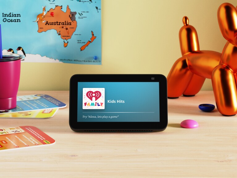 An image of an Amazon Echo Show 5 device that reads "Kids Hits" and "Try 'Alexa, lets play a game'." The background shows a gold toy in the shape of a dog, a poster of the map of Australia, and assorted game cards.