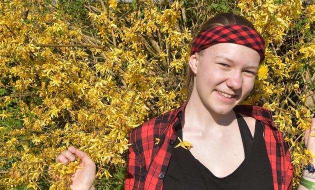 Ein Mädchen mit rotem Haarband steht vor einer Blumenhecke und schaut in die Kamera. Sie lächelt und sieht fröhlich aus.
