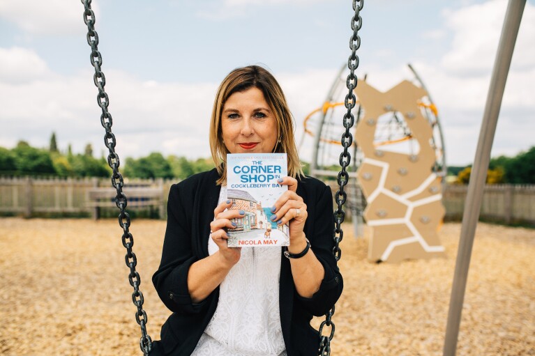 KDP author Nicola May sitting on a swing, holding here newest novel: The Corner Shop in Cockleberry Bay