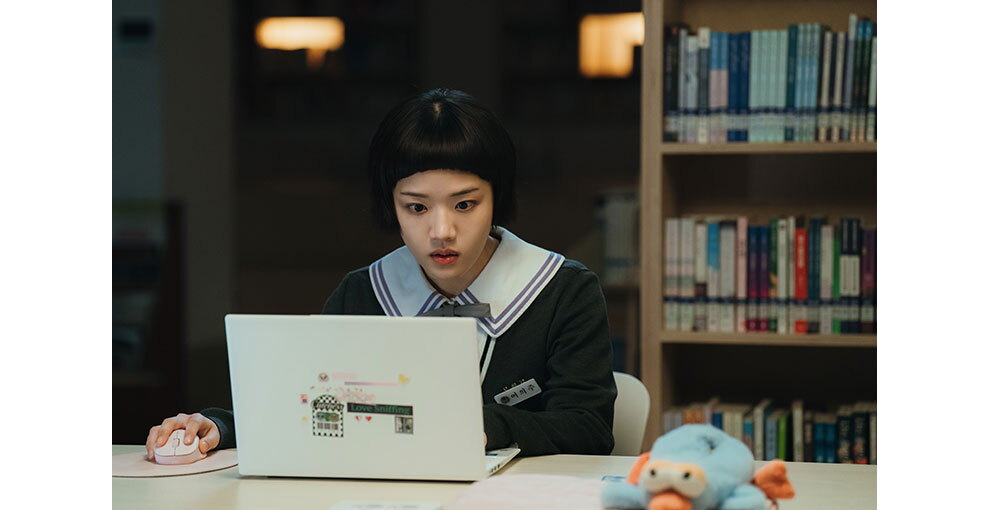図書室で机に向かい、ノートパソコンの画面を見つめる少女。襟の大きな制服を着用。机の右側にぬいぐるみ。