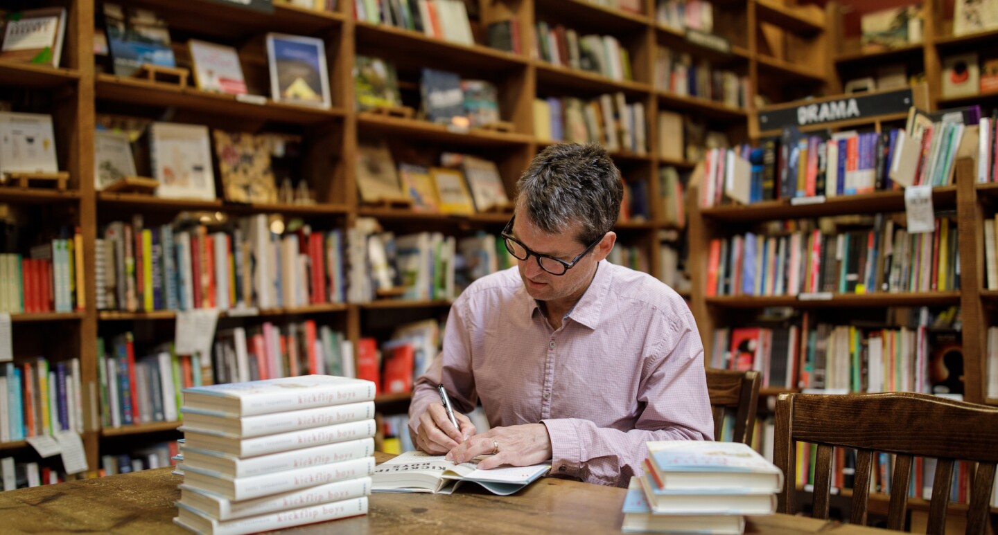 An author sits at a table and signs his book in a bookstore, surrounded by shelves of other book titles.