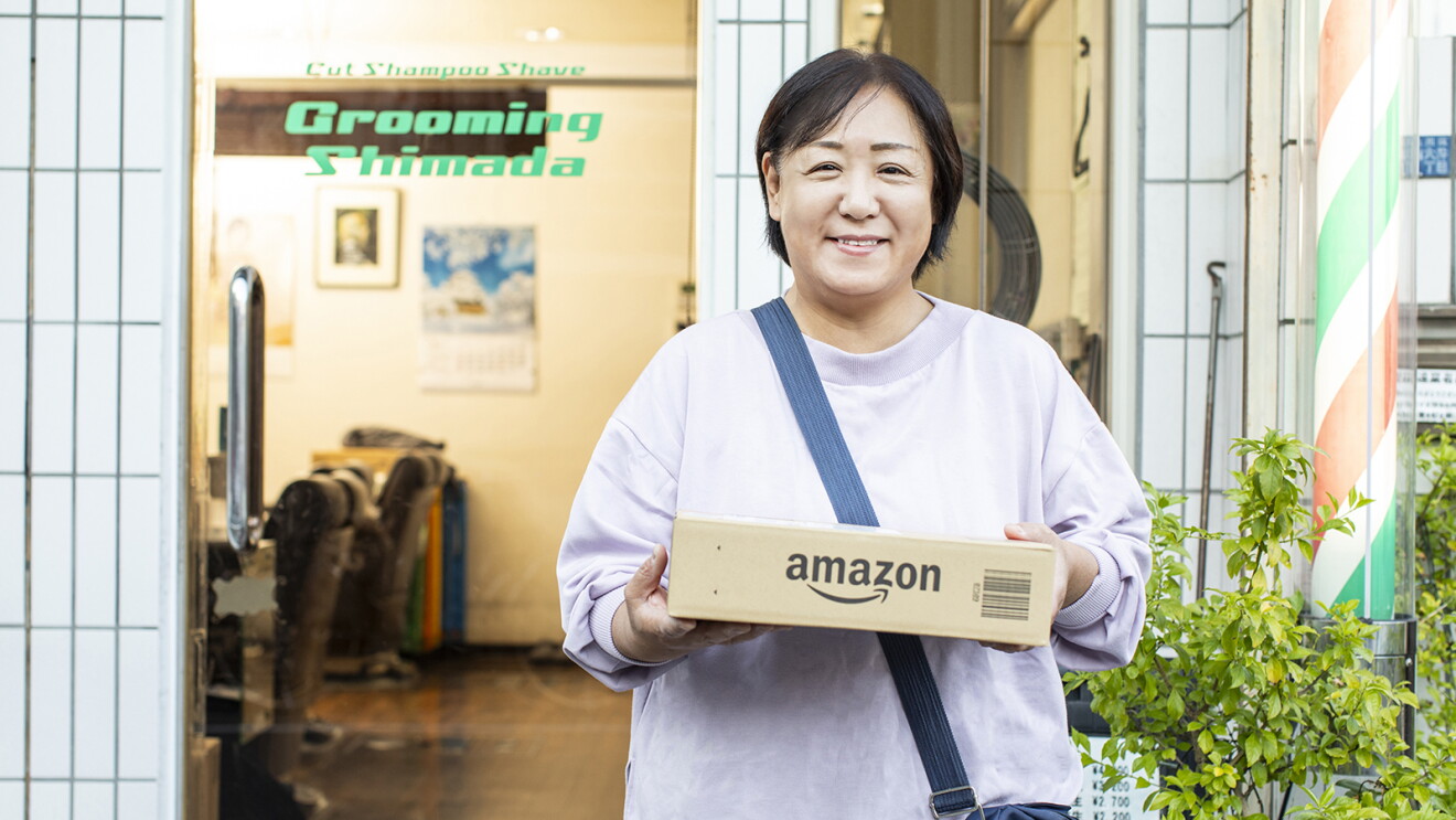 理容店の前に立ち、Amazonのロゴ入り段ボール箱を両手で持つ女性。背後に理容店のポールや植木が見える。
