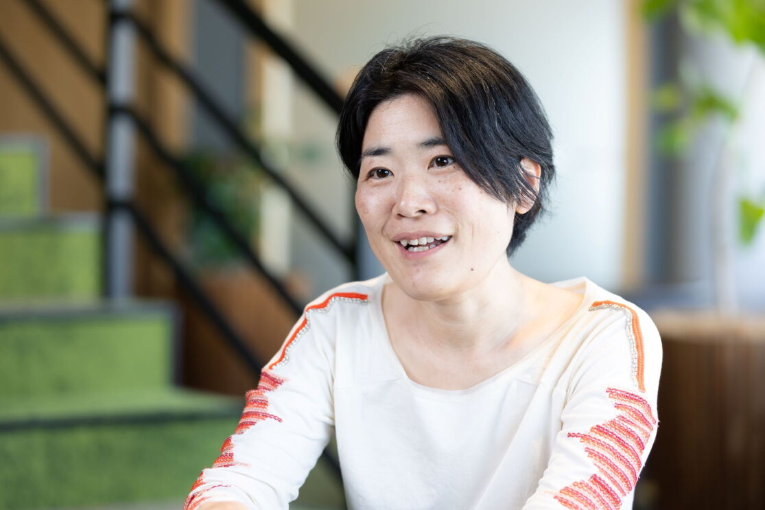 白い長そでのシャツを着ている女性が話している。彼女の後ろには緑色の階段と観葉植物が見える