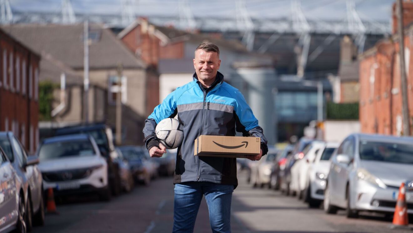 Shay Given in an Amazon-branded jacket is holding a package and soccer ball on residential street