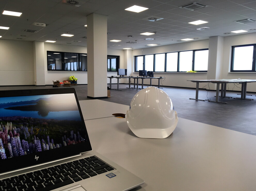 Auf einem weißen Schreibtisch steht links ein Laptop, rechts ein weißer Bauhelm. Im Hintergrund Schreibtische mit einigen Bildschirmen. Die Bürofläche ist noch nicht fertig eingerichtet.