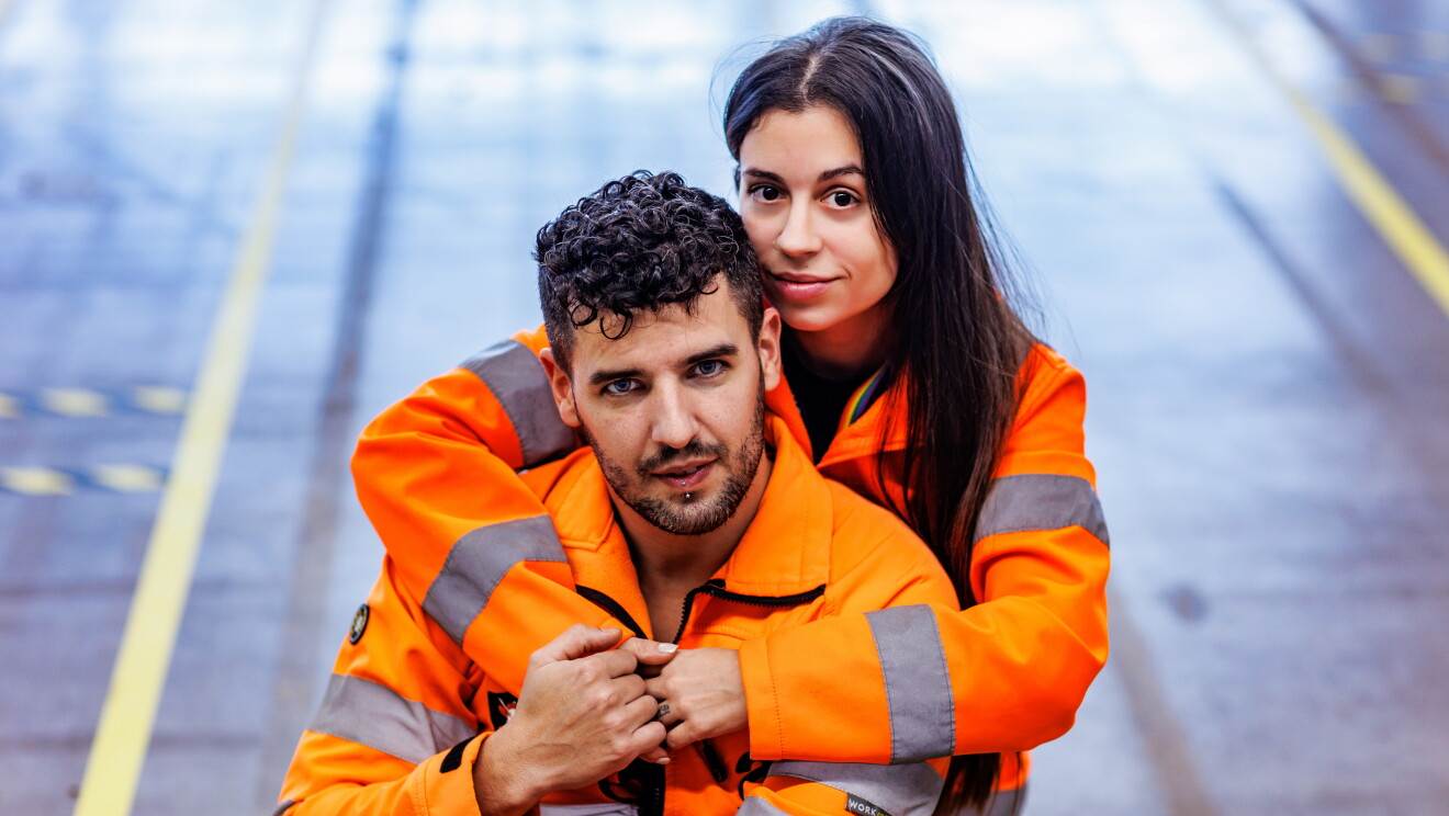 Miriam y Mario en una foto artística sacada desde arriba donde se les ve mirando a cámara y agarrados. Ella por detrás le abraza y él le agarra una de las manos. Los dos van con una chaqueta naranja que simula un chaleco de seguridad. De fondo el pasillo de un centro logístico. Miriam lleva el pelo largo por la cintura y es de color castaño. Él tiene el pelo corto pero la parte superior la lleva un poco más larga y la tiene rizada. Tiene los ojos claro, barba de dos días y un piercing en el labio inferior.
