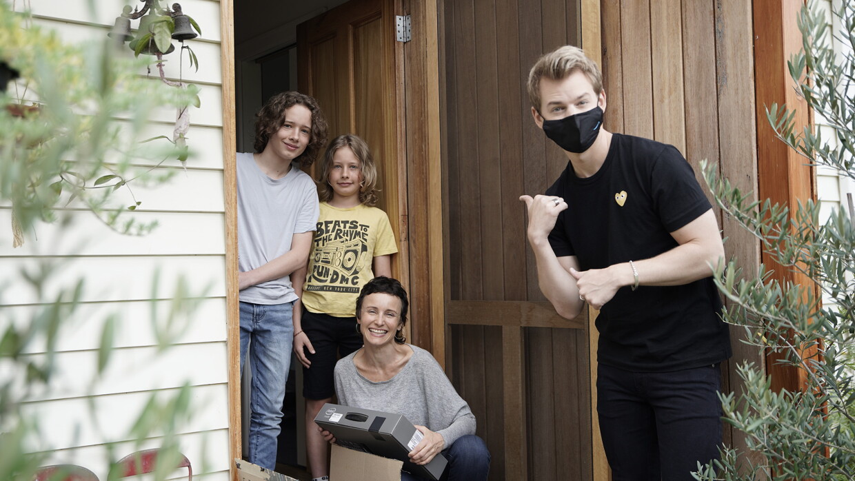 A man stands by the front door of a home, he wears a black mask and black outfit, looking at the camera, he has his hand in a gesture that suggests "look at that / them," toward a family smiling at the camera. 