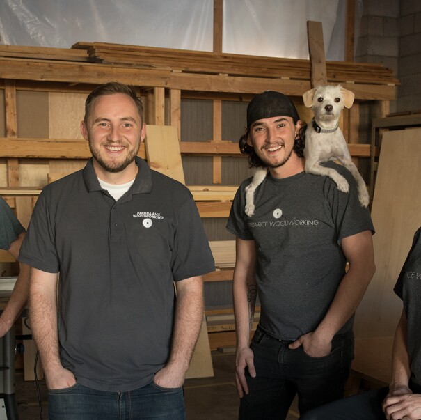Three males and a female stand in front of a wall of wood. One of the men has a dog perched on his shoulders. These four individuals are siblings who run a small business named Pereida-Rice Woodworking, which sells on Amazon.