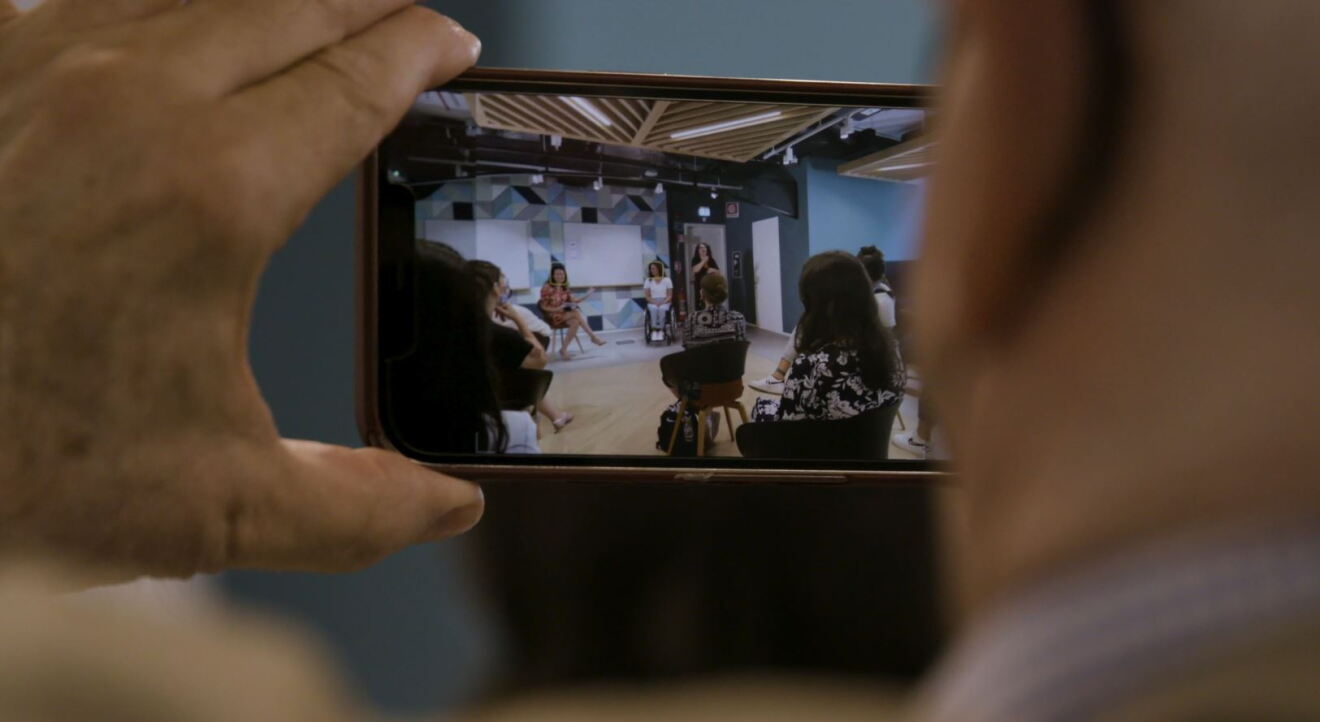 Primo piano delle mani di un uomo che reggono uno smartphone con la fotocamera attiva. Sta riprendendo 3 donne sul fondo di una sala. Una è Giulia Ghiretti, è in sedia a rotelle. 